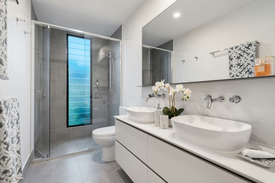 A white bathroom with twin basins, a toilet and a glass shower
